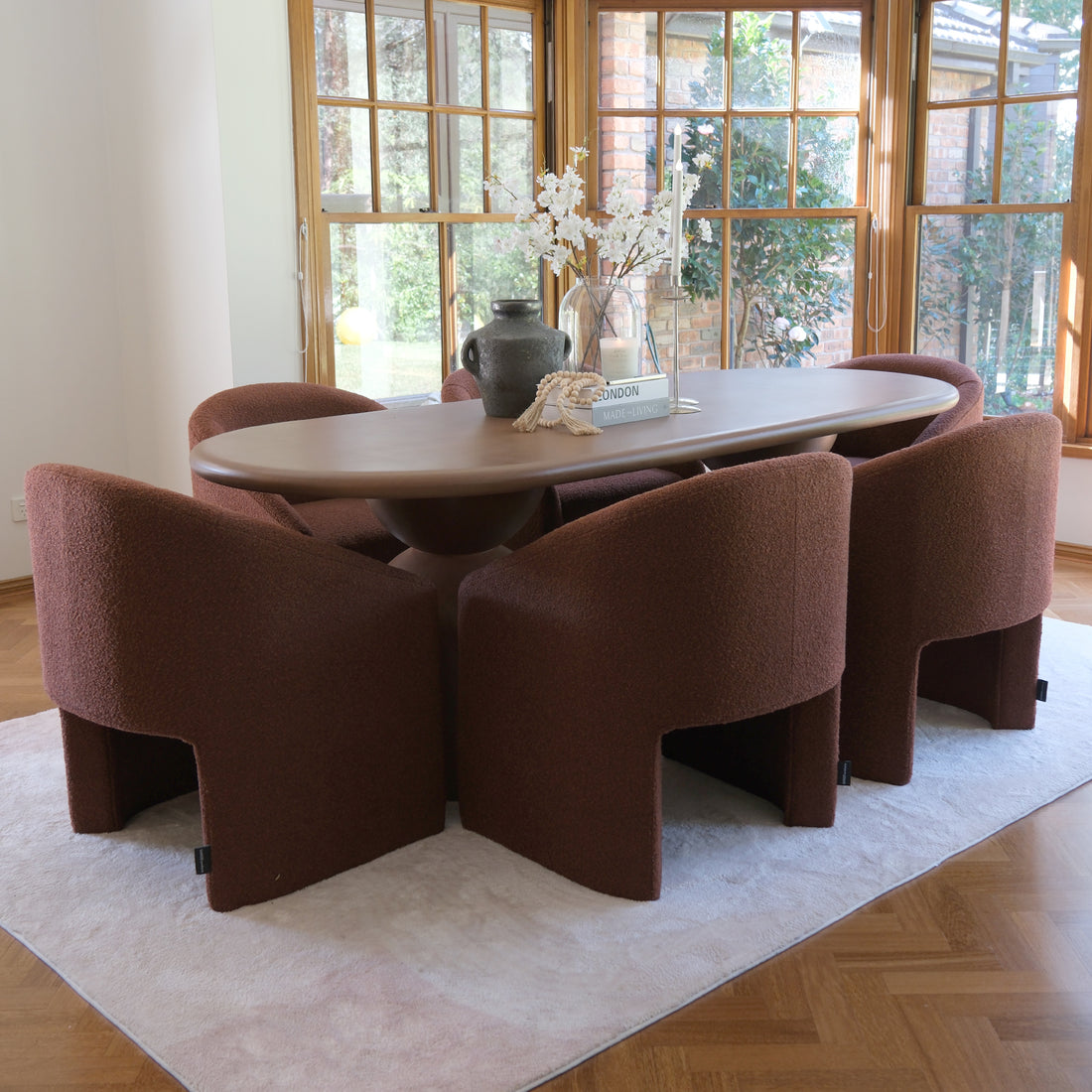 Nuve Bouclé Upholstered Accent Armchair in Rust Brown, featured in a cozy dining nook, showcases its curved design and rich texture amidst elegant vases and candles on an oval wooden table.