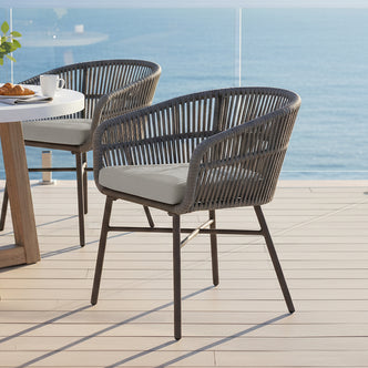 Marula Rope Outdoor Dining Chairs in Brown Set of 4, featuring woven-rope armchairs with metal legs and light cushions, beside a round table with coffee and croissants, overlooking a tranquil ocean view.