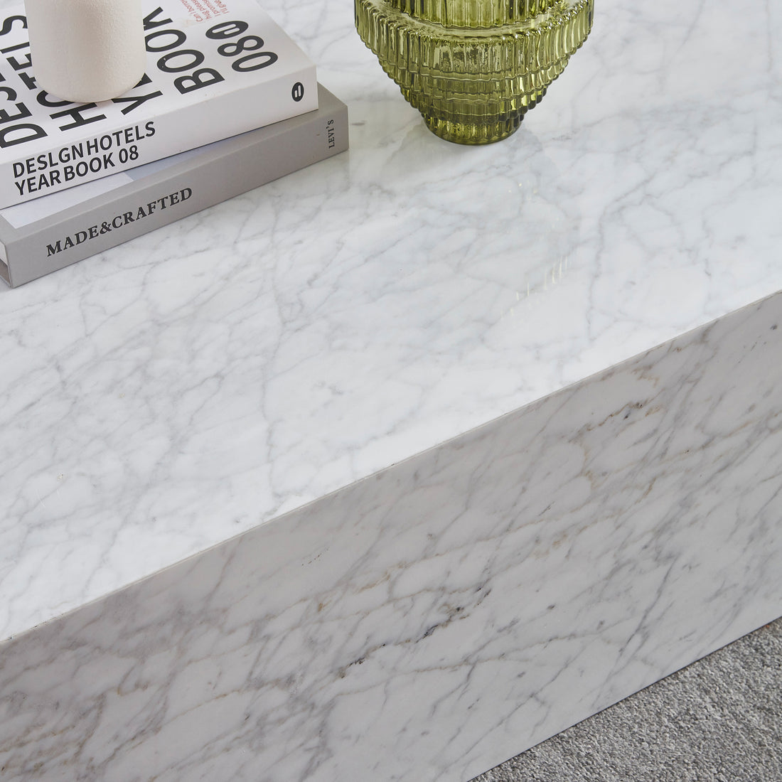 Solene 100cm Coffee Table in Carrara with stacked design books, white ceramic holder, and green ribbed vase atop its marble surface, resting on a light grey carpet.