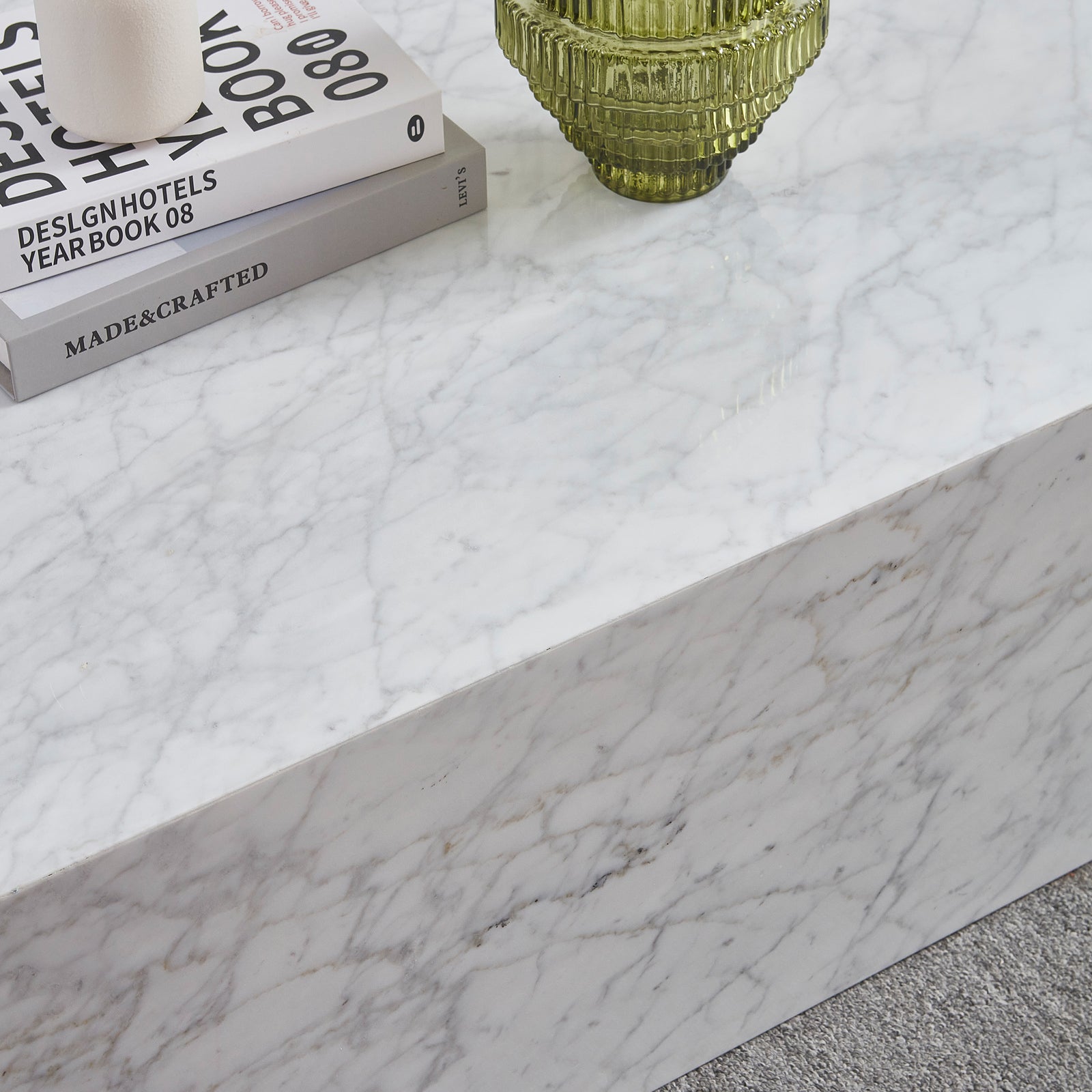 Solene 100cm Coffee Table in Carrara with stacked design books, white ceramic holder, and green ribbed vase atop its marble surface, resting on a light grey carpet.