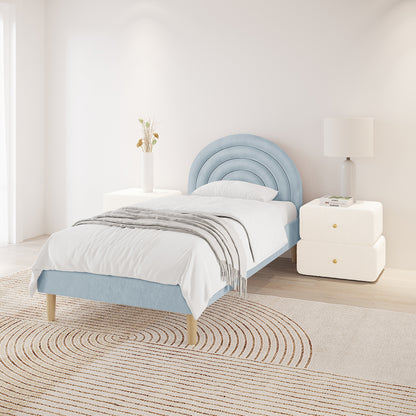Estilla Velvet Rainbow Bed in Ice Blue features a semi-circular upholstered headboard, white bedding, and light gray throw, flanked by cream nightstands with a vase and lamp, atop a neutral rug.