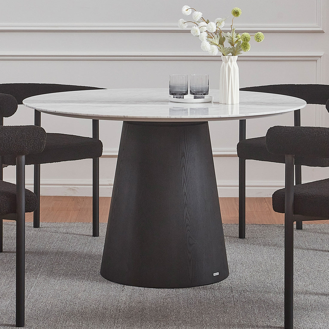 Dina 120cm Round Marble Dining Table in Carrara Black features a white marble top and black wooden base, surrounded by four textured black chairs. A white ceramic tray and vase adorn the table.