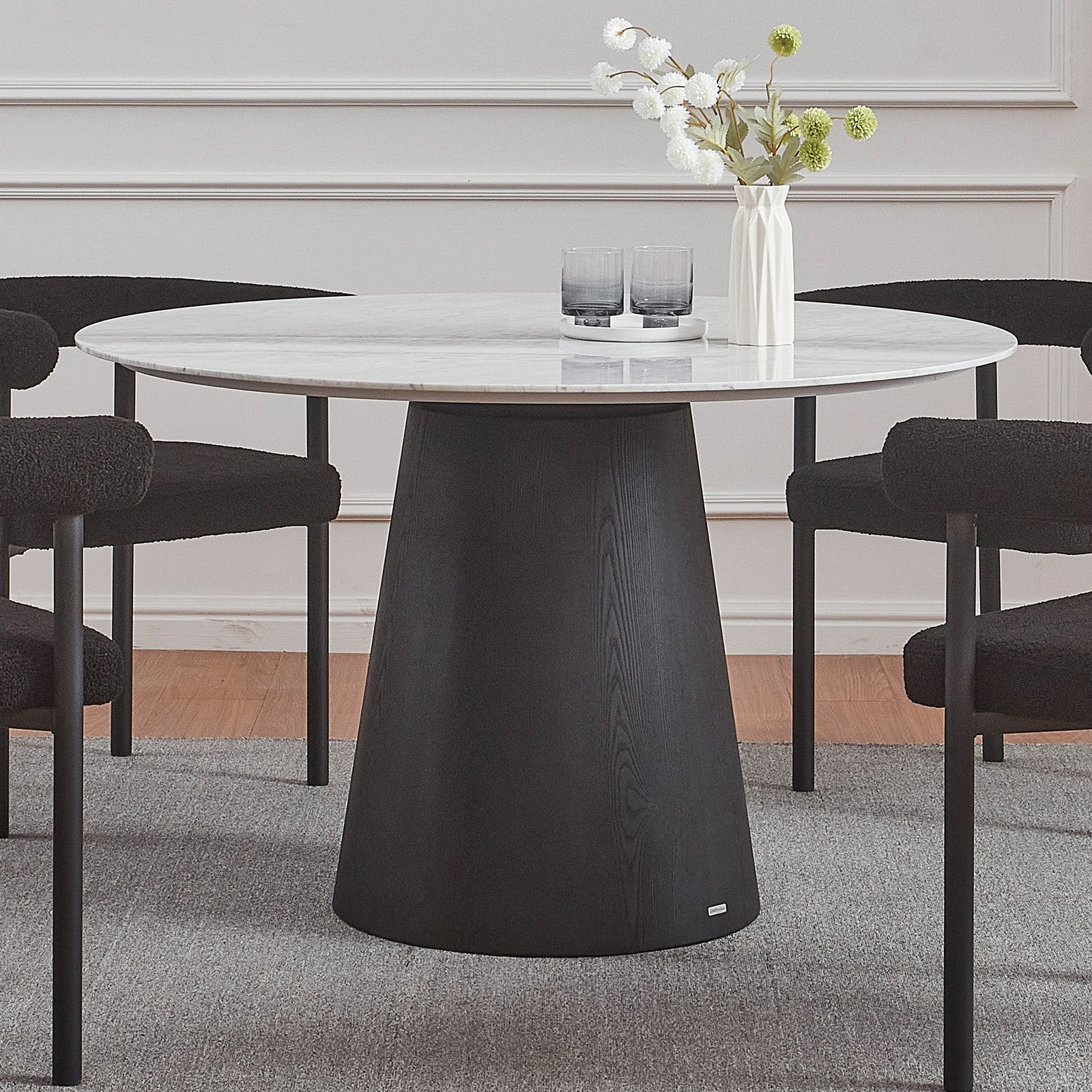 Dina 120cm Round Marble Dining Table in Carrara Black features a white marble top and black wooden base, surrounded by four textured black chairs. A white ceramic tray and vase adorn the table.