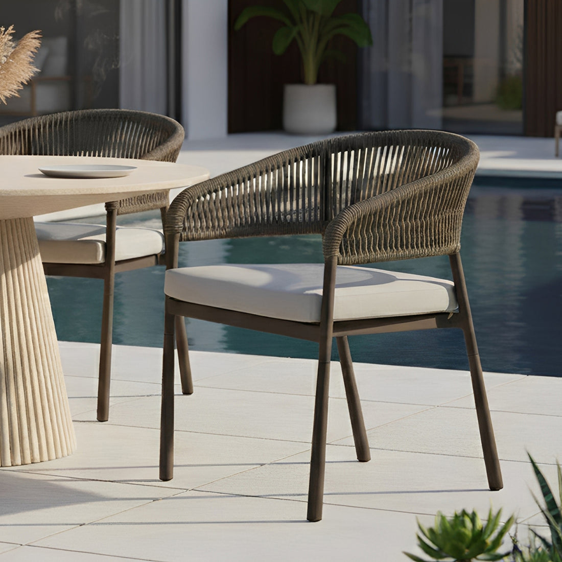 Coastal Outdoor Rope Dining Chairs Set of 2 in Brown, featuring dark metal frames and woven rope backs with light seat cushions, situated in a modern poolside setting.