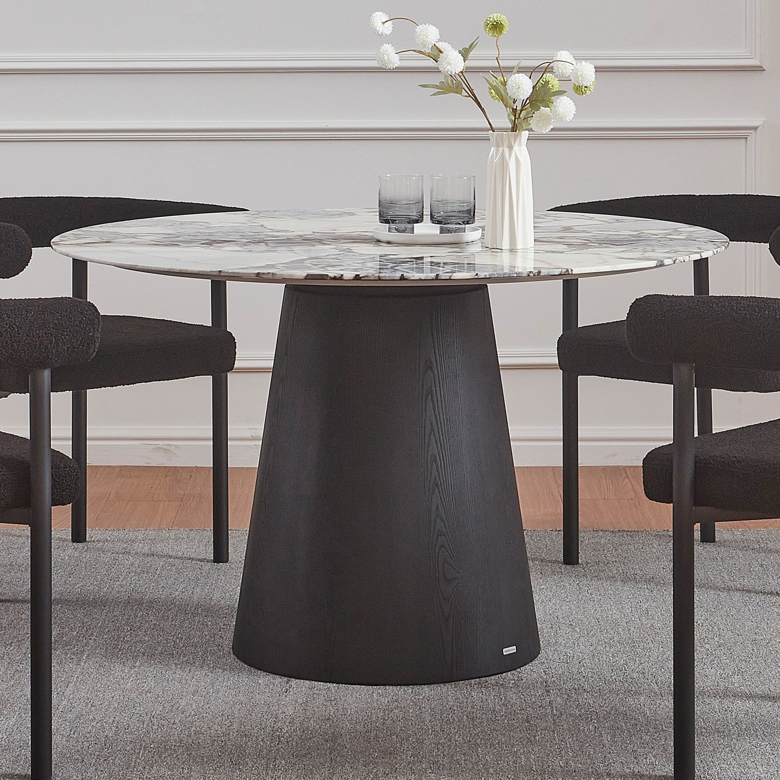 Dina 120cm Round Marble Dining Table in Blue Galaxy Black features a white-grey marble top with veining and a black tapered pedestal, surrounded by black boucle-style chairs with metal legs.