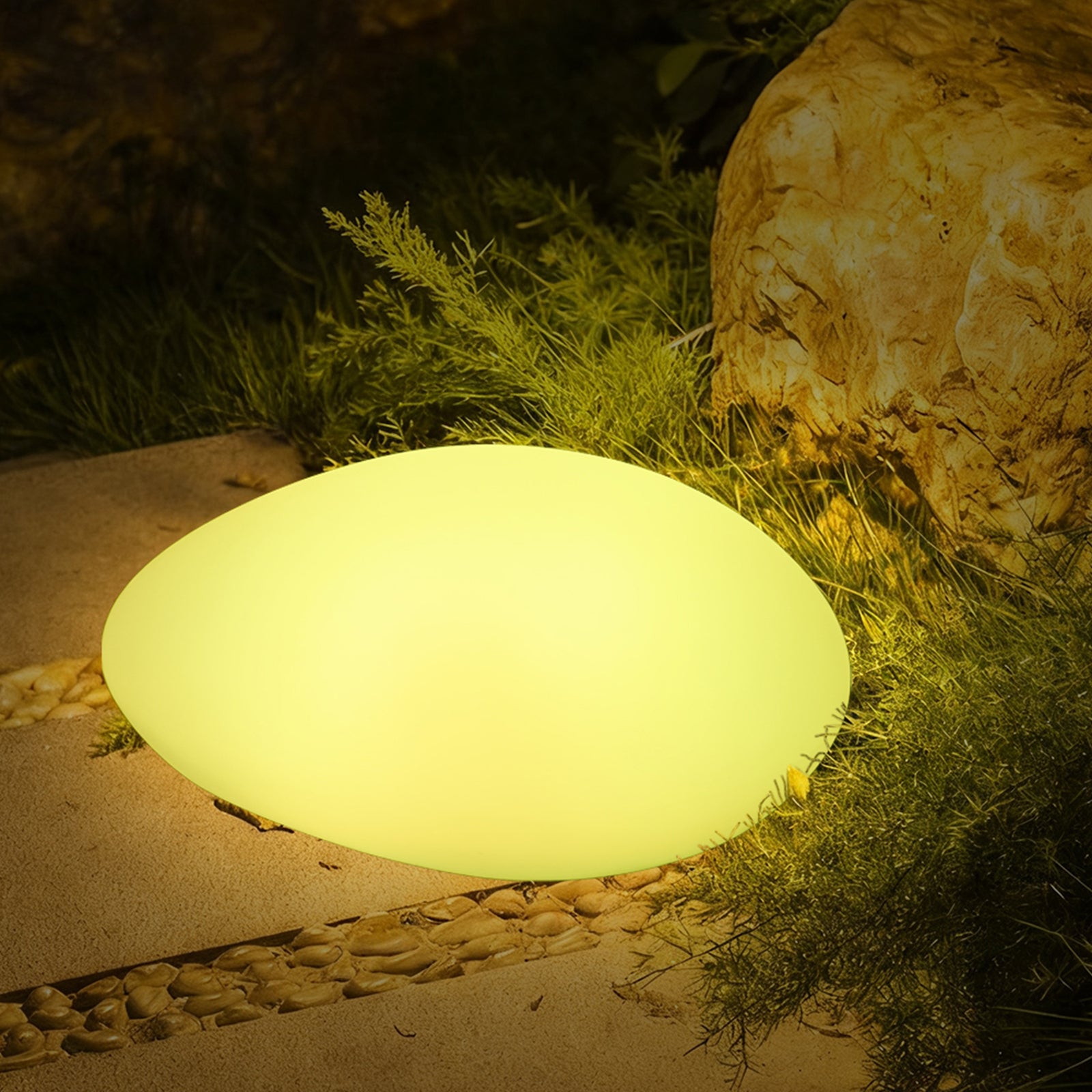 Solar Charging Outdoor LED Rock Light on a garden path, mimicking a natural stone, emits a soft glow beside greenery and a larger boulder, enhancing outdoor ambiance.