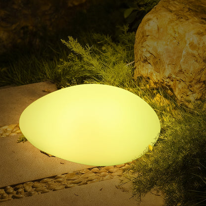 Solar Charging Outdoor LED Rock Light on a garden path, mimicking a natural stone, emits a soft glow beside greenery and a larger boulder, enhancing outdoor ambiance.