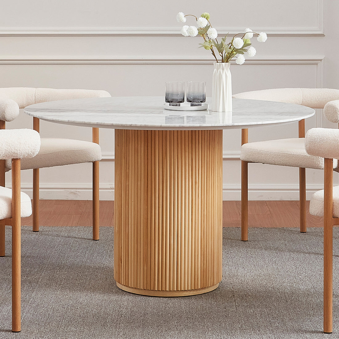 Gracie 120cm Round Marble Dining Table in Natural with a white marble top, fluted oak base, surrounded by cream bouclé chairs, features a ceramic vase and tray with glasses on top.