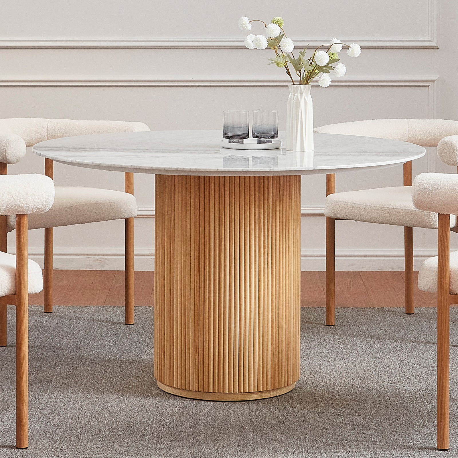Gracie 120cm Round Marble Dining Table in Natural with a white marble top, fluted oak base, surrounded by cream bouclé chairs, features a ceramic vase and tray with glasses on top.