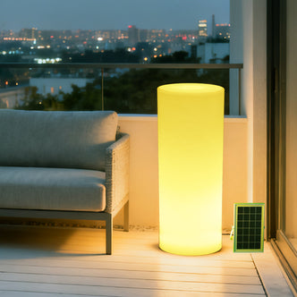 70cm Outdoor Solar LED Bollard Light on a modern balcony, glowing warmly beside a gray-upholstered sofa, with a solar panel leaning against the wall, city skyline visible.
