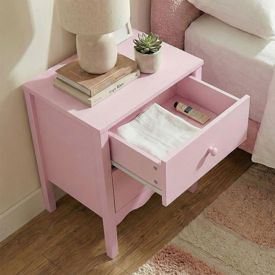 Pastel pink nightstand in a cozy bedroom with lamp, books, plant, and open drawer