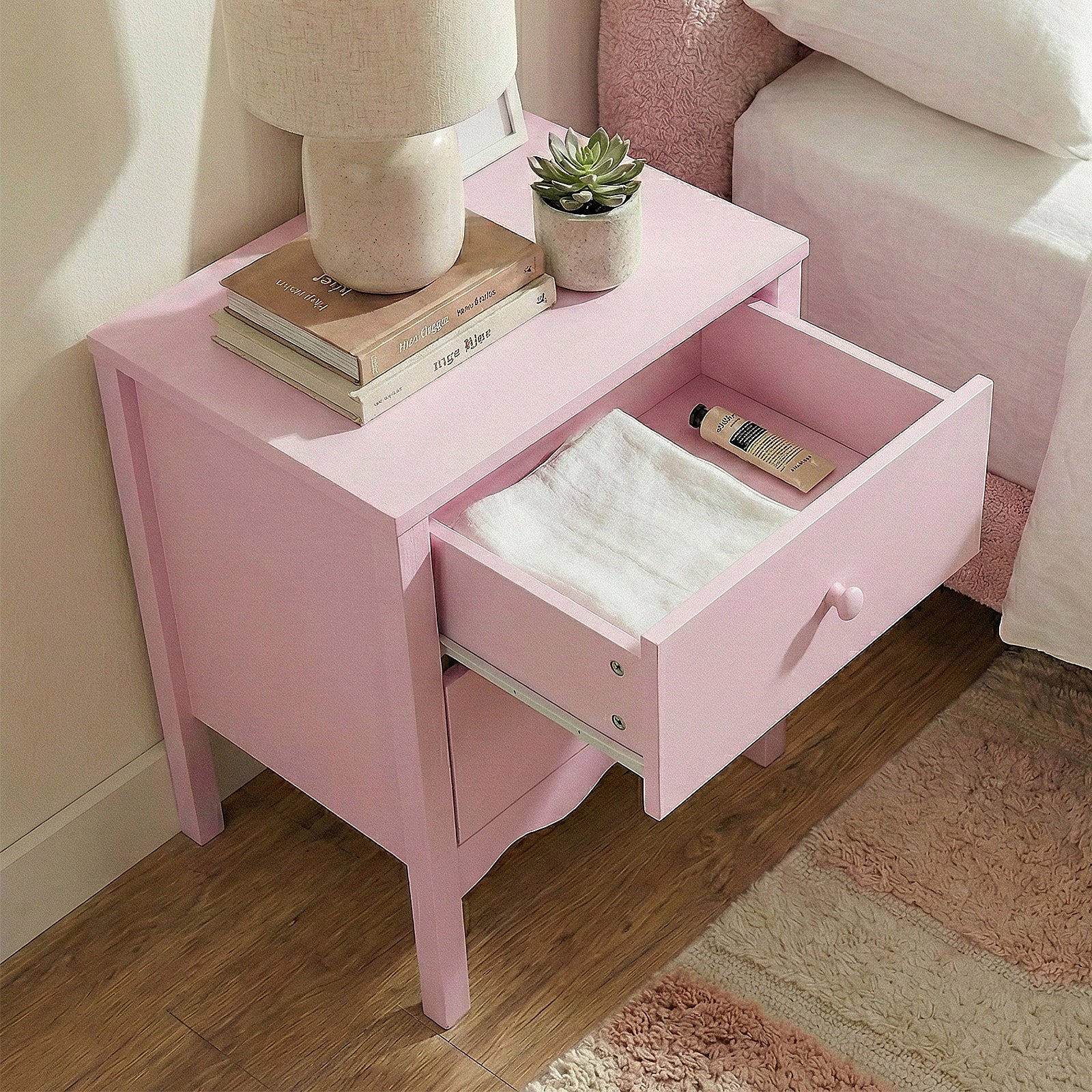 Pastel pink nightstand in a cozy bedroom with lamp, books, plant, and open drawer