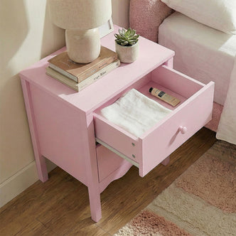Pastel pink nightstand in a cozy bedroom with lamp, books, plant, and open drawer