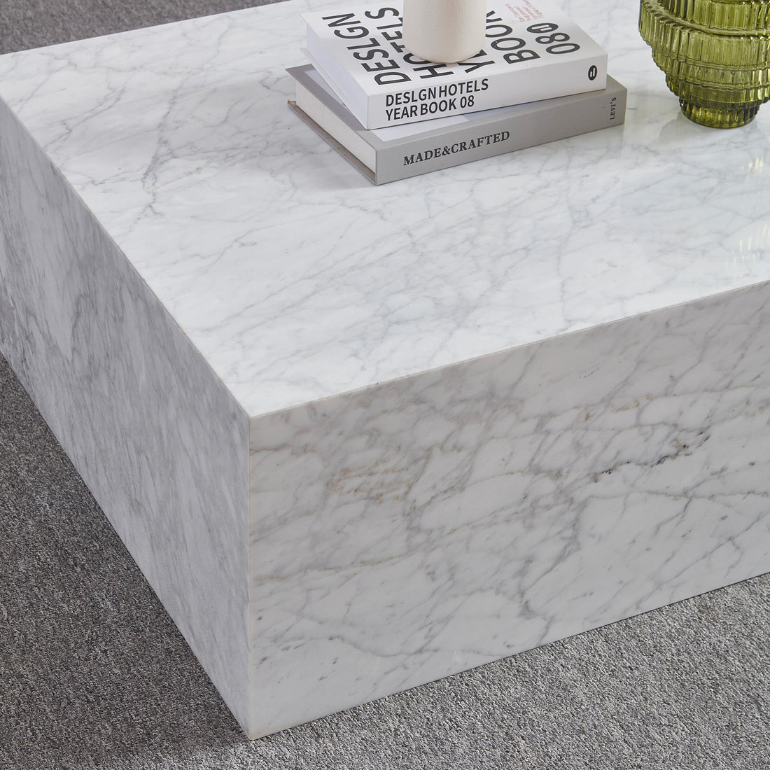 Solene 100cm Coffee Table in Carrara, a low rectangular marble piece, features two stacked books and a cylindrical object on top, set on a grey loop-pile carpet.