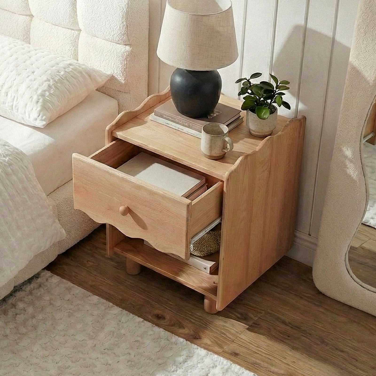 Wooden nightstand next to a bed in a lifestyle setting with lamp, books, and plant