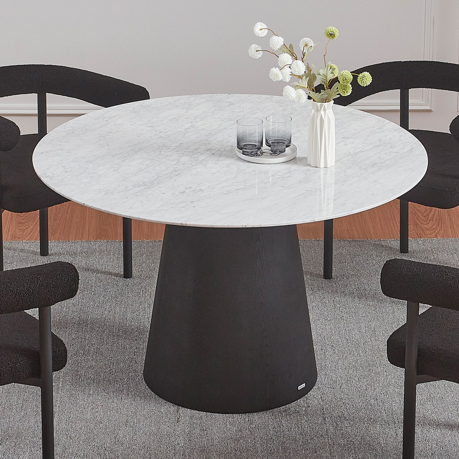 Dina 120cm Round Marble Dining Table in Carrara Black, featuring a white marble top and black pedestal base, surrounded by four low-back black chairs on a light gray rug, with decorative accents.
