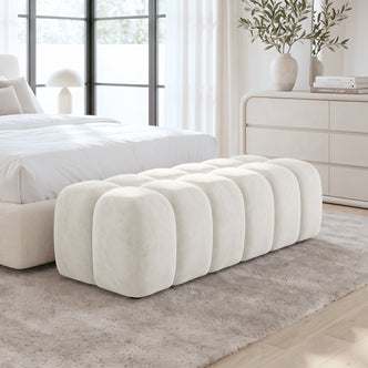 Light beige upholstered ottoman bench at the foot of a bed in a bright bedroom