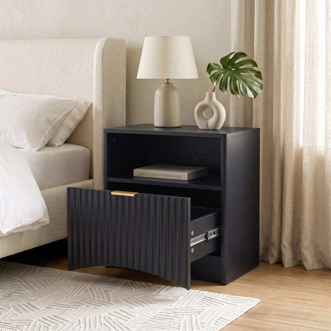 Black nightstand with open drawer in a styled bedroom next to a bed and lamp