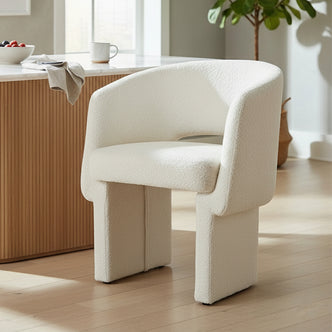 Avela Bouclé Fabric Curved Accent Armchair in Cream White, featuring a high back and armrests, sits near a light wood counter with a mug, bowl of berries, and cloth napkin.