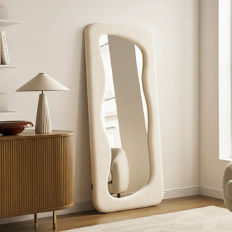 Sabrina 180cm Curved Full Length Mirror in Beige features a padded, undulating frame against a pale wall, enhancing a minimalist living room corner with a serene, contemporary feel.