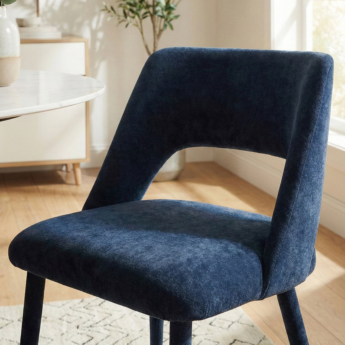 Dark blue velvet chair at edge of round table on a patterned rug, natural room setting