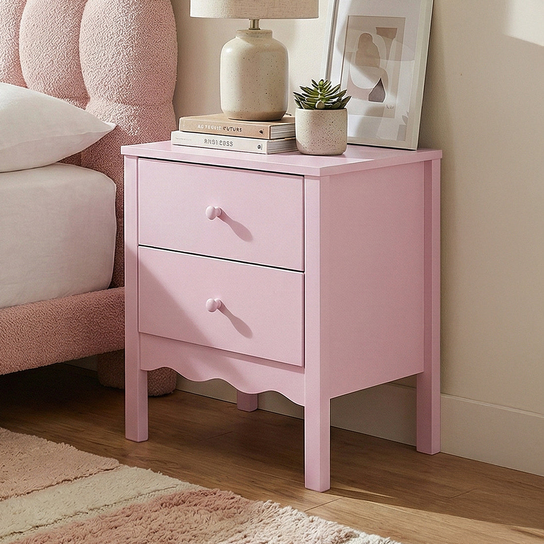 Pink two-drawer nightstand styled next to a bed with lamp, books, and plant in a furnished room
