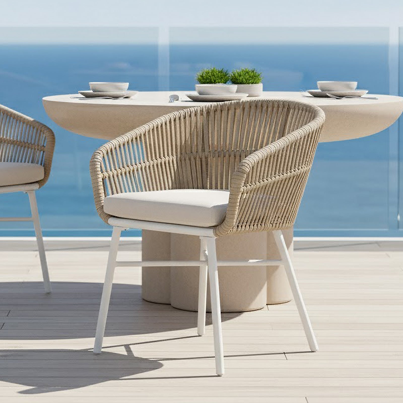 Marula Rope Outdoor Dining Chairs in White Set of 4 with woven rope design, cushioned seats, and slim metal legs on a light-wood deck, paired with a round table overlooking the sea.