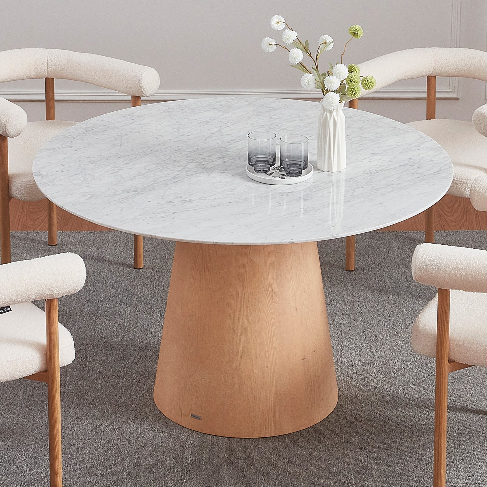 Dina 120cm Round Marble Dining Table in Carrara Natural with a white marble top and light wood pedestal base, surrounded by four cream chairs, white ceramic tray, and a tall vase with pom-pom flowers.