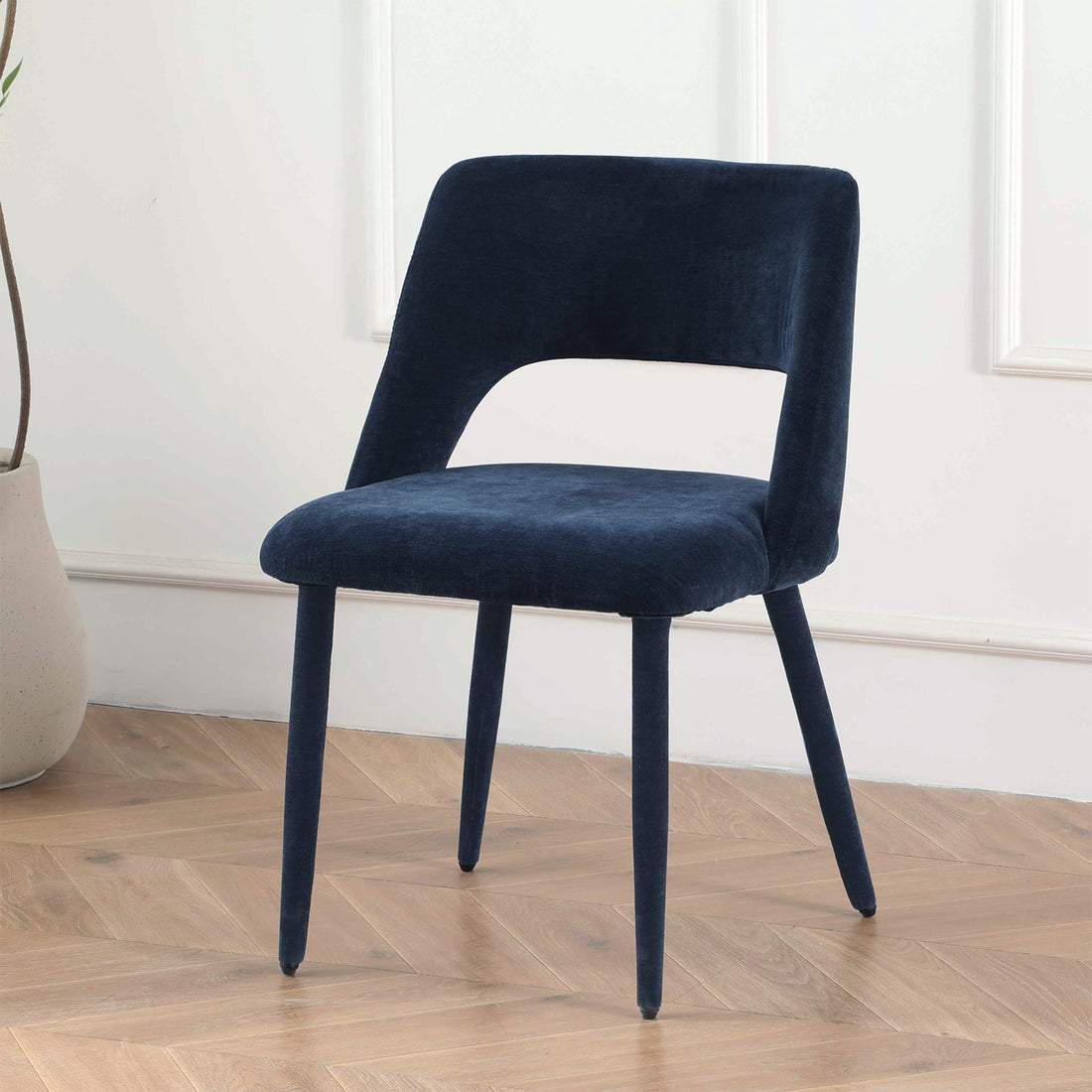 Dark blue upholstered chair on herringbone wood floor in front of white paneled wall with decorative vase
