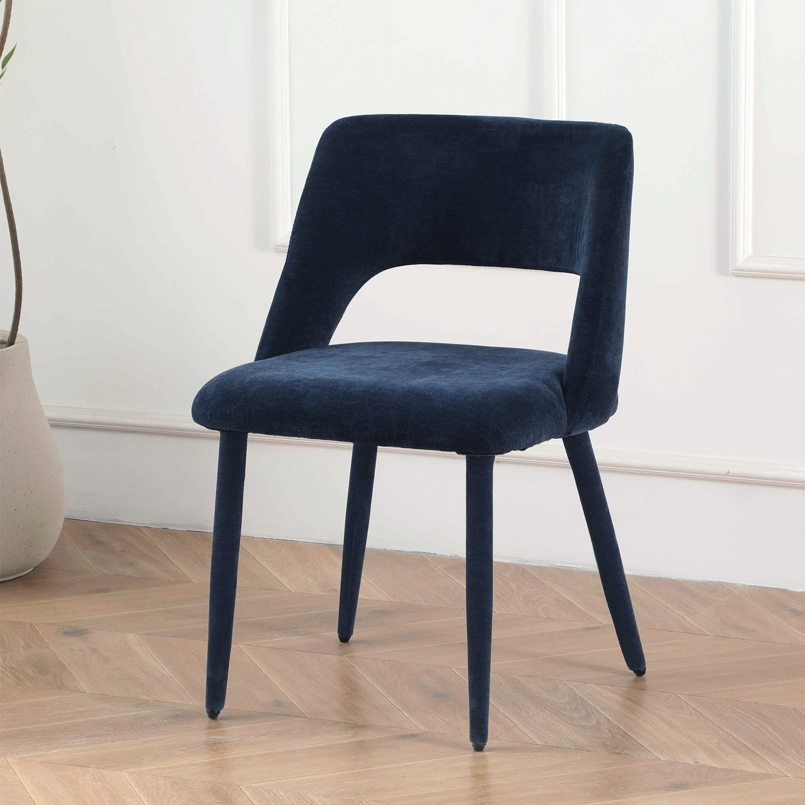 Dark blue upholstered chair on herringbone wood floor in front of white paneled wall with decorative vase