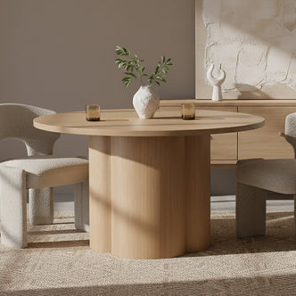 Cece 120cm Round Wooden Dining Table in Oak with scalloped pedestal base, white vase with green branches, amber glasses on top, flanked by cream bouclé chairs on natural-fiber rug.