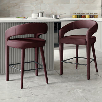 Eisha Velvet Bar Stool Set of 2 in Dark Plum features upholstered stools with rounded backs, padded seats, and slim wooden legs with metal footrests, complementing a contemporary kitchen or breakfast bar setting.
