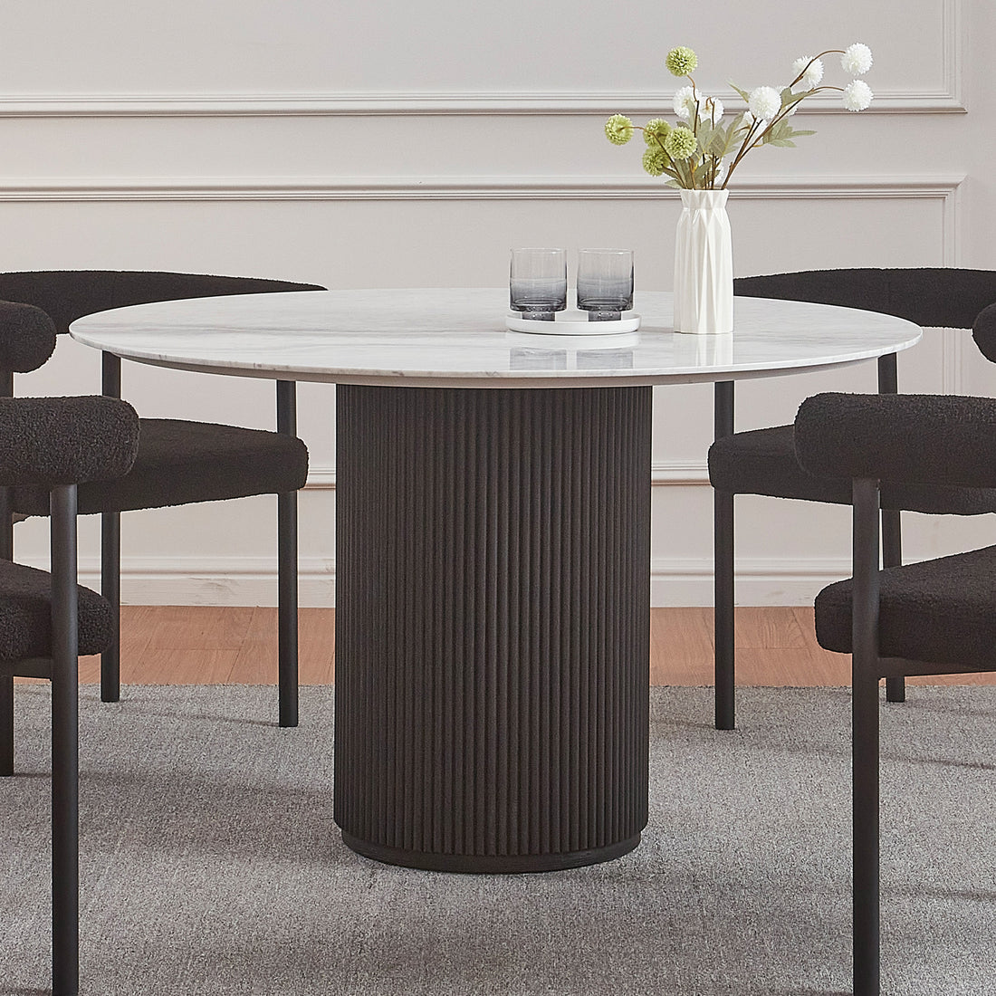 Gracie 120cm Round Marble Dining Table in Black features a white marble top on a black fluted base, surrounded by four plush black chairs, complemented by a vase and glasses on top.