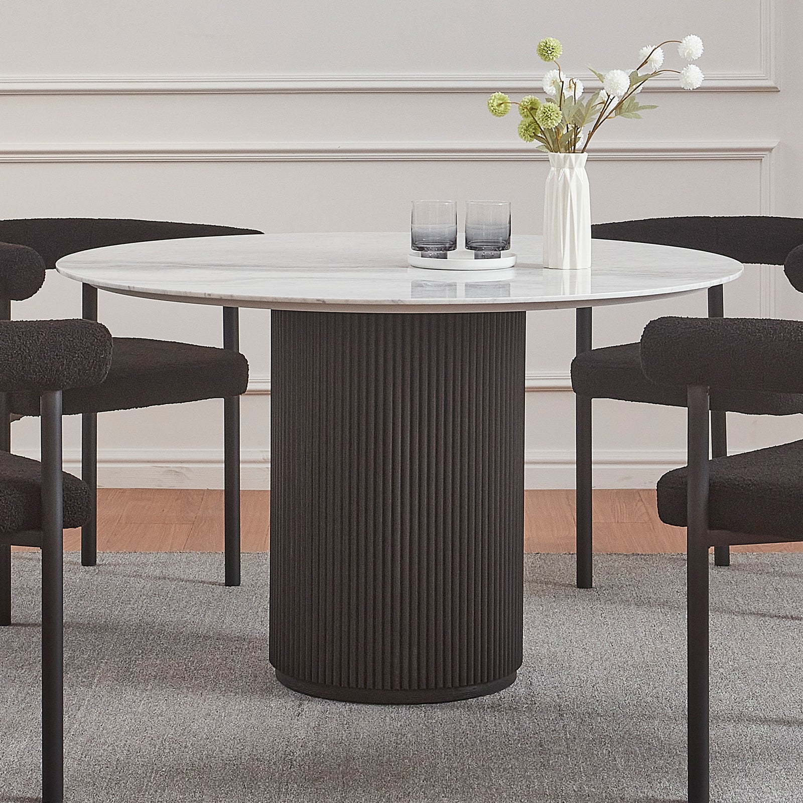 Gracie 120cm Round Marble Dining Table in Black features a white marble top on a black fluted base, surrounded by four plush black chairs, complemented by a vase and glasses on top.