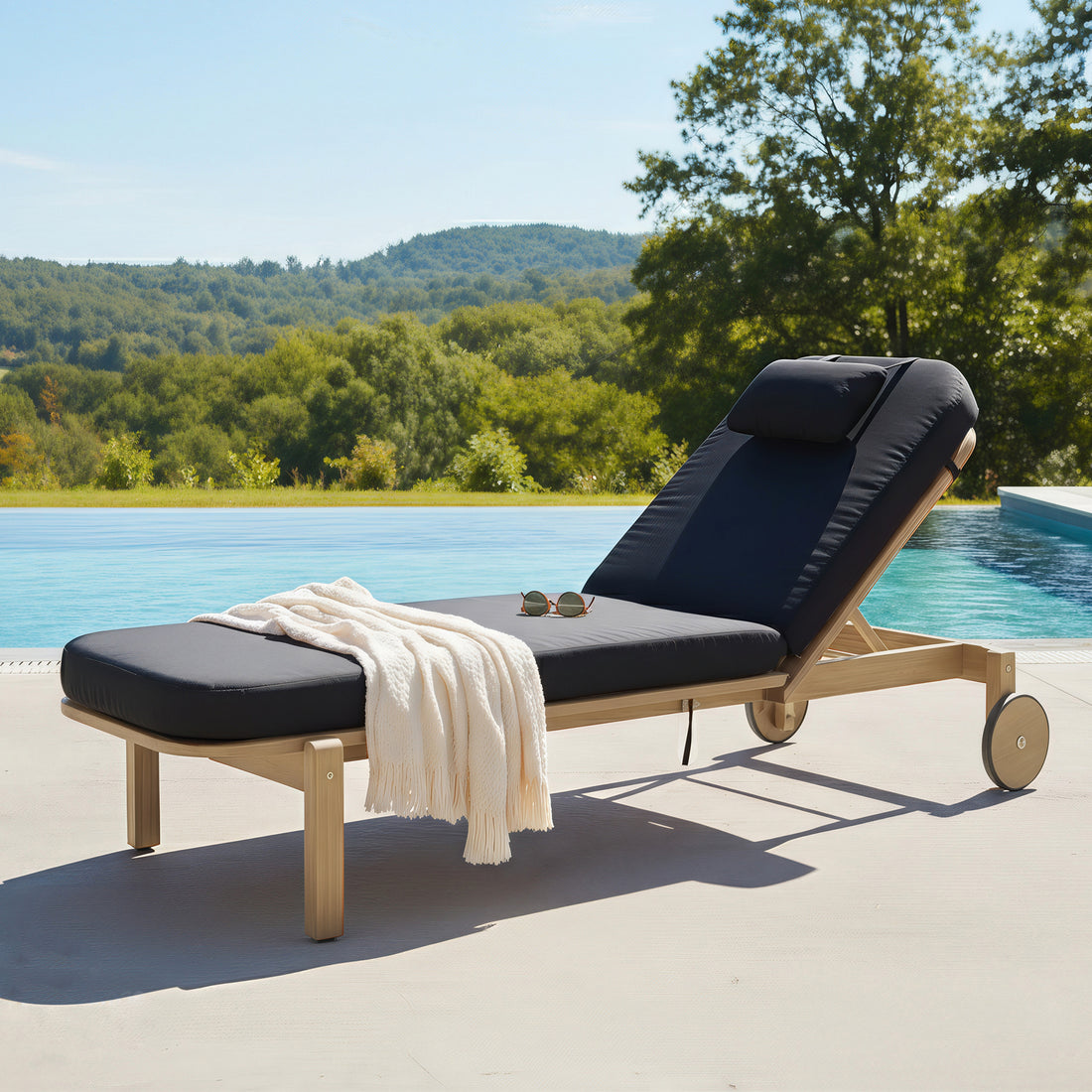 Zyra Hardwood Reclining Sun Lounger with Cushion in Black on poolside deck, featuring light-wood frame, adjustable backrest, cream throw, and sunglasses. Blue pool and scenic hills in background.