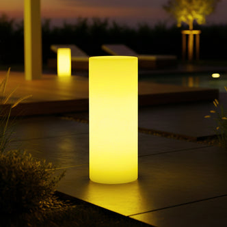 16 Colour Outdoor LED Bollard Light in Multicolour glowing warmly on a tiled patio, with a second lamp on a wooden deck, set against a backdrop of lounge chairs and a softly lit tree.