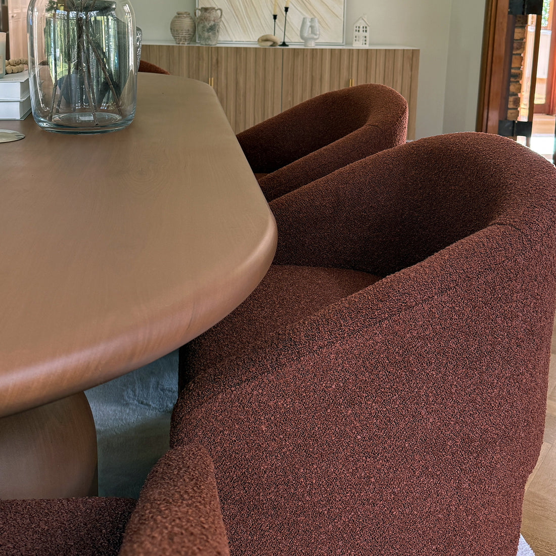 Nuve Bouclé Upholstered Accent Armchair in Rust Brown, featuring a sculpted silhouette and deep rust-brown boucle fabric, is tucked around a wooden table with a vase of twigs.