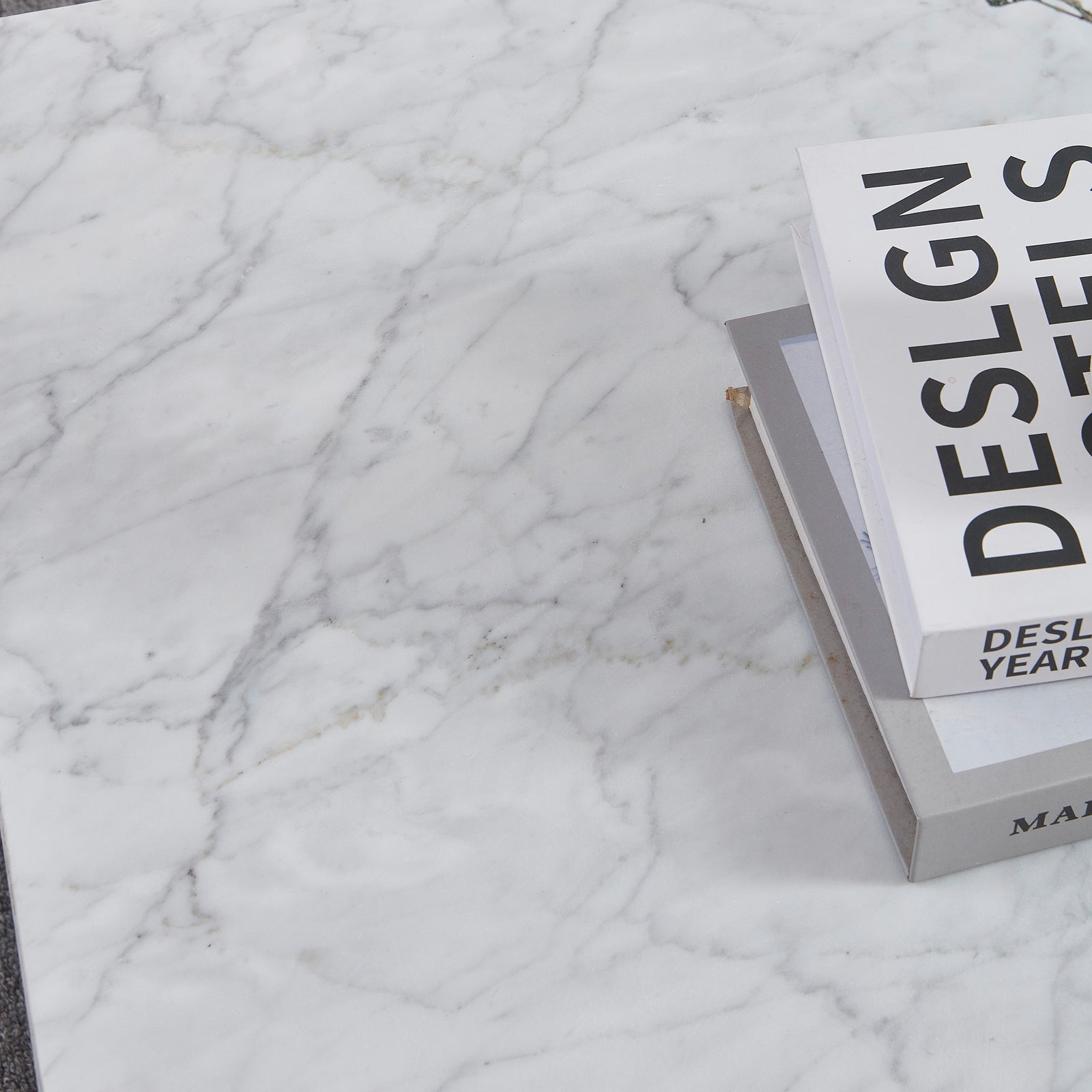 Solene 100cm Coffee Table in Carrara on a white marble surface, featuring a small stack of hardcover books. The table showcases its elegant, authentic marble veining and sleek, low-profile design.