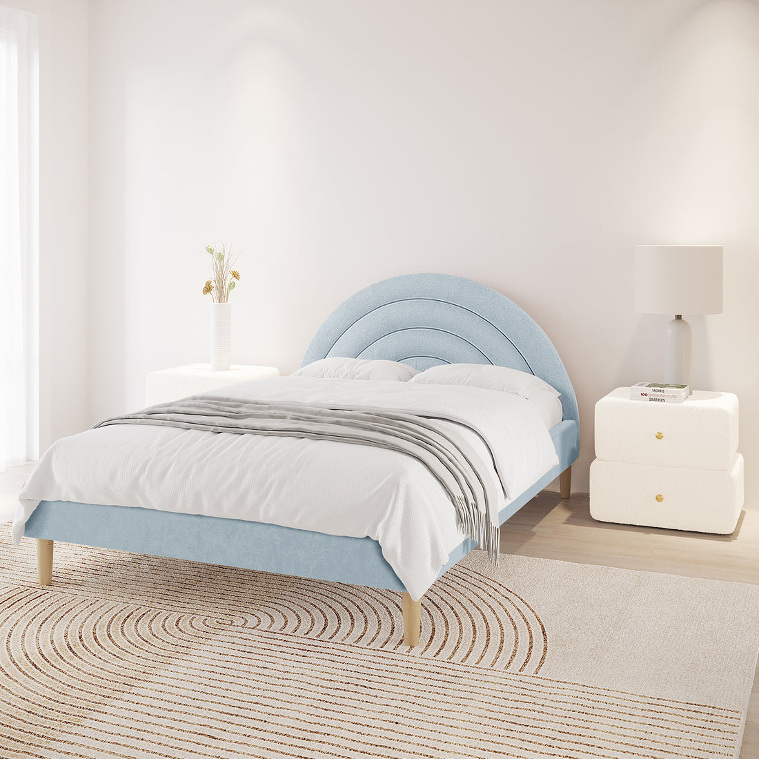 Estilla Velvet Rainbow Bed in Ice Blue, featuring a concentric semi-circle headboard, white linens, gray throw, and adjacent white side tables with a vase and lamp, in a sunlit, minimalist bedroom.