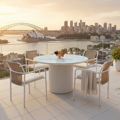Evone 4 Seater Stone Outdoor Dining Table Set featuring a round marble top with four rope-back chairs, glass bowl of fruit, and wine glasses, overlooking a harbor at sunset.