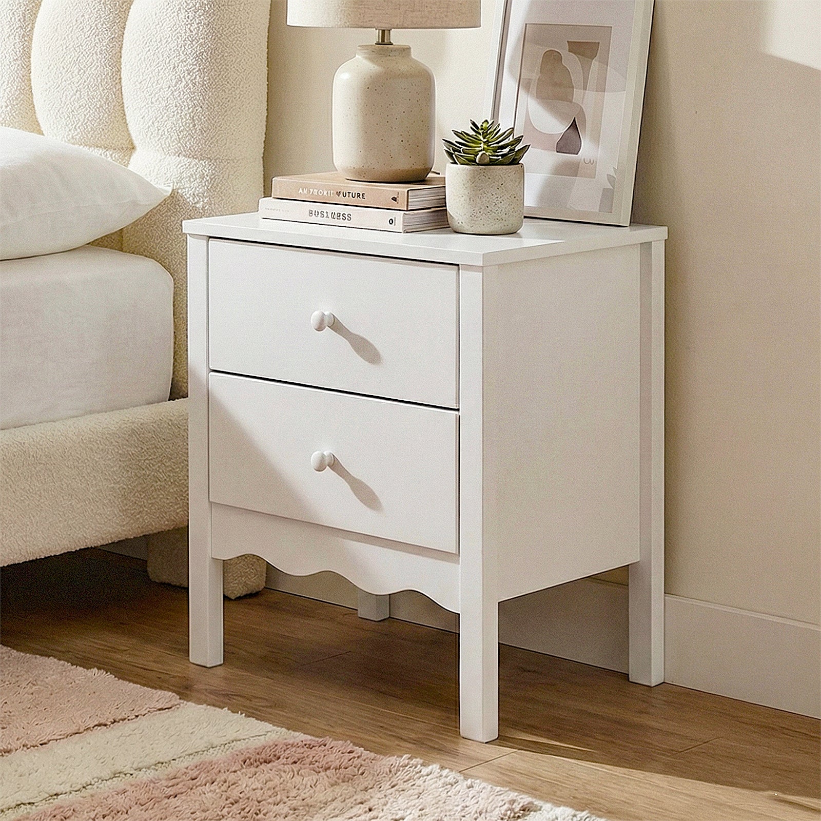 White two-drawer nightstand styled next to a bed with lamp, books, and plant in a bedroom