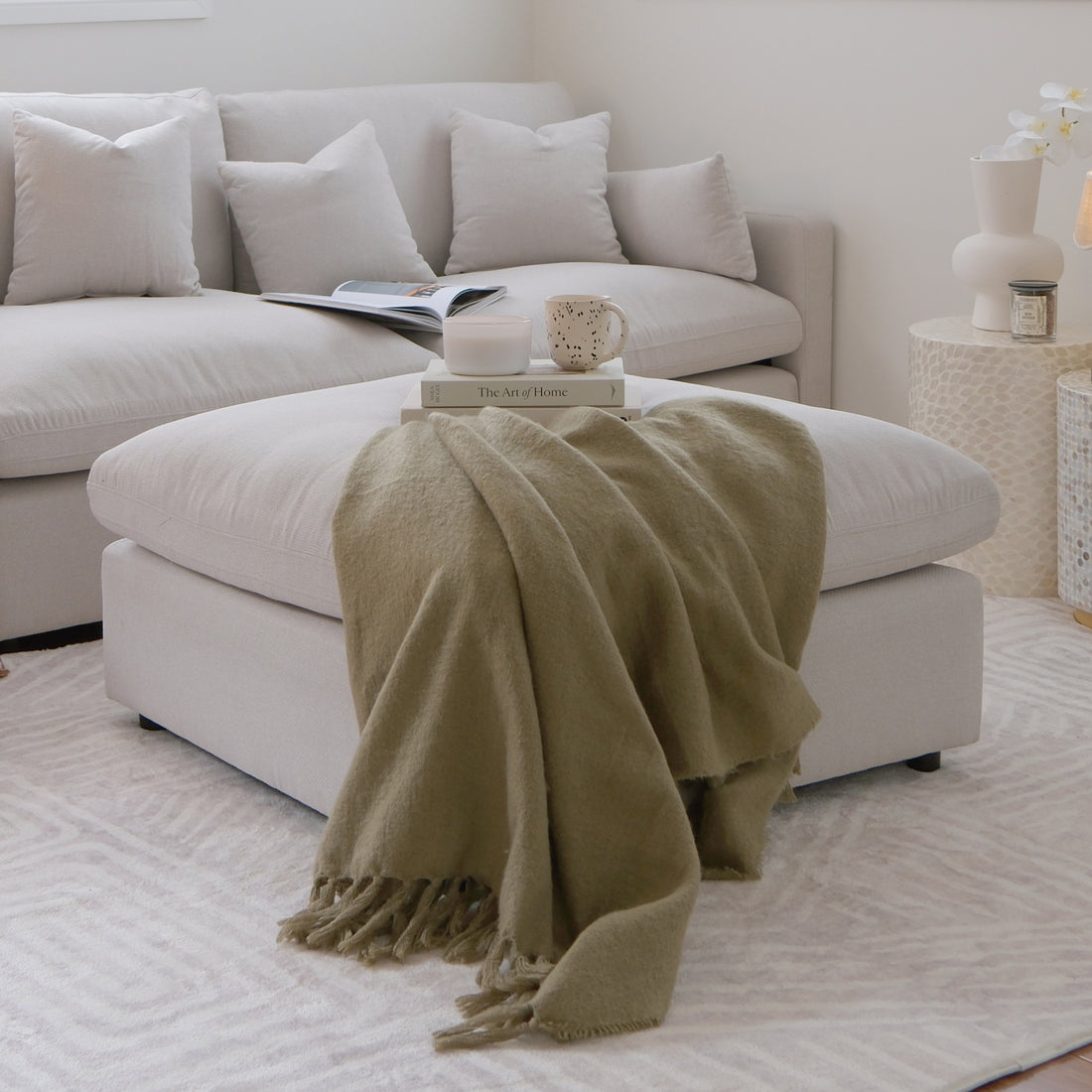 Light beige ottoman styled in a modern living room with sectional sofa, books, and decor
