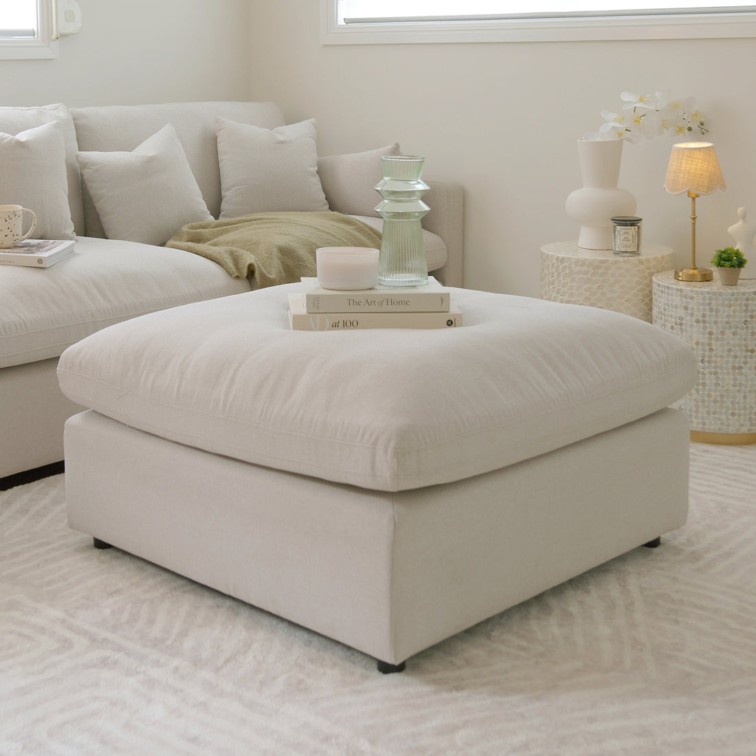 Light beige square ottoman styled in a modern living room with books, candles, and decor.
