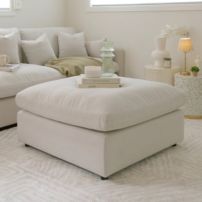 Light beige square ottoman styled in a modern living room with books, candles, and decor.