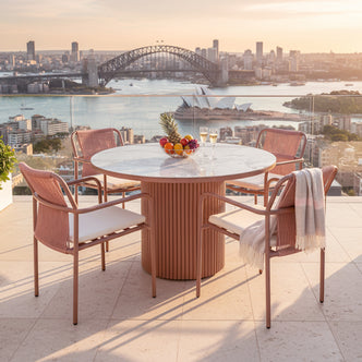 Evone 4 Seater Stone Outdoor Dining Table Set in Terracotta features a marble top and four rope-style armchairs. A bowl of fruit and champagne flutes enhance the sunlit rooftop ambiance.