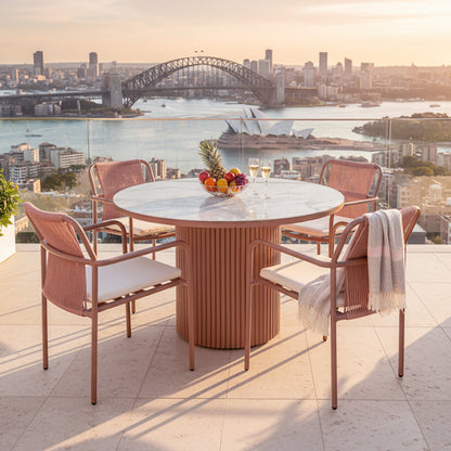 Evone 4 Seater Stone Outdoor Dining Table Set in Terracotta features a marble top and four rope-style armchairs. A bowl of fruit and champagne flutes enhance the sunlit rooftop ambiance.