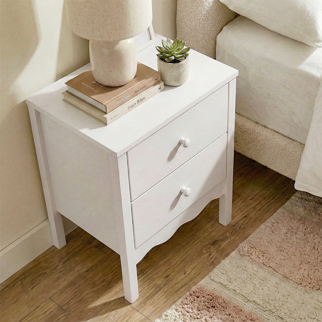White two-drawer nightstand next to bed with lamp, books, and plant on top in a cozy room