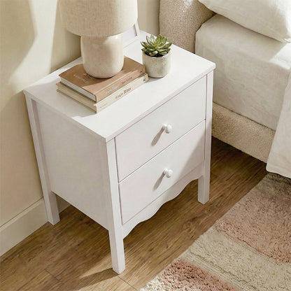 White two-drawer nightstand next to bed with lamp, books, and plant on top in a cozy room