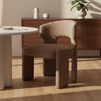 Lunari Bouclé Curved Accent Chair in Brown, upholstered in warm beige boucle fabric, stands on a light wood floor with a high backrest, near a wooden sideboard with ceramics and a plant.