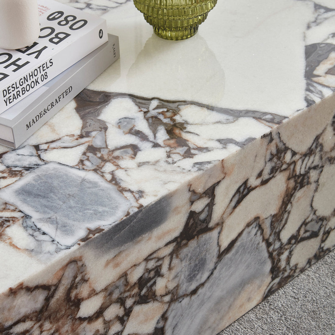 Solene 100cm Coffee Table in Blue Galaxy, a polished marble piece with cream, gray, and rust veining. Topped with design books and a green textured glass vessel, on a light carpeted floor.